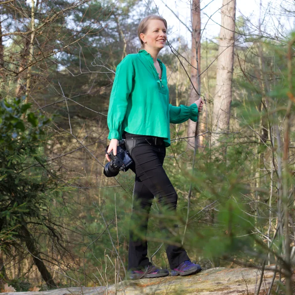 natuurfotograaf en verkoopster van wanddecoraties uit Limburg staat met haar camera in het bos op een boomstam.